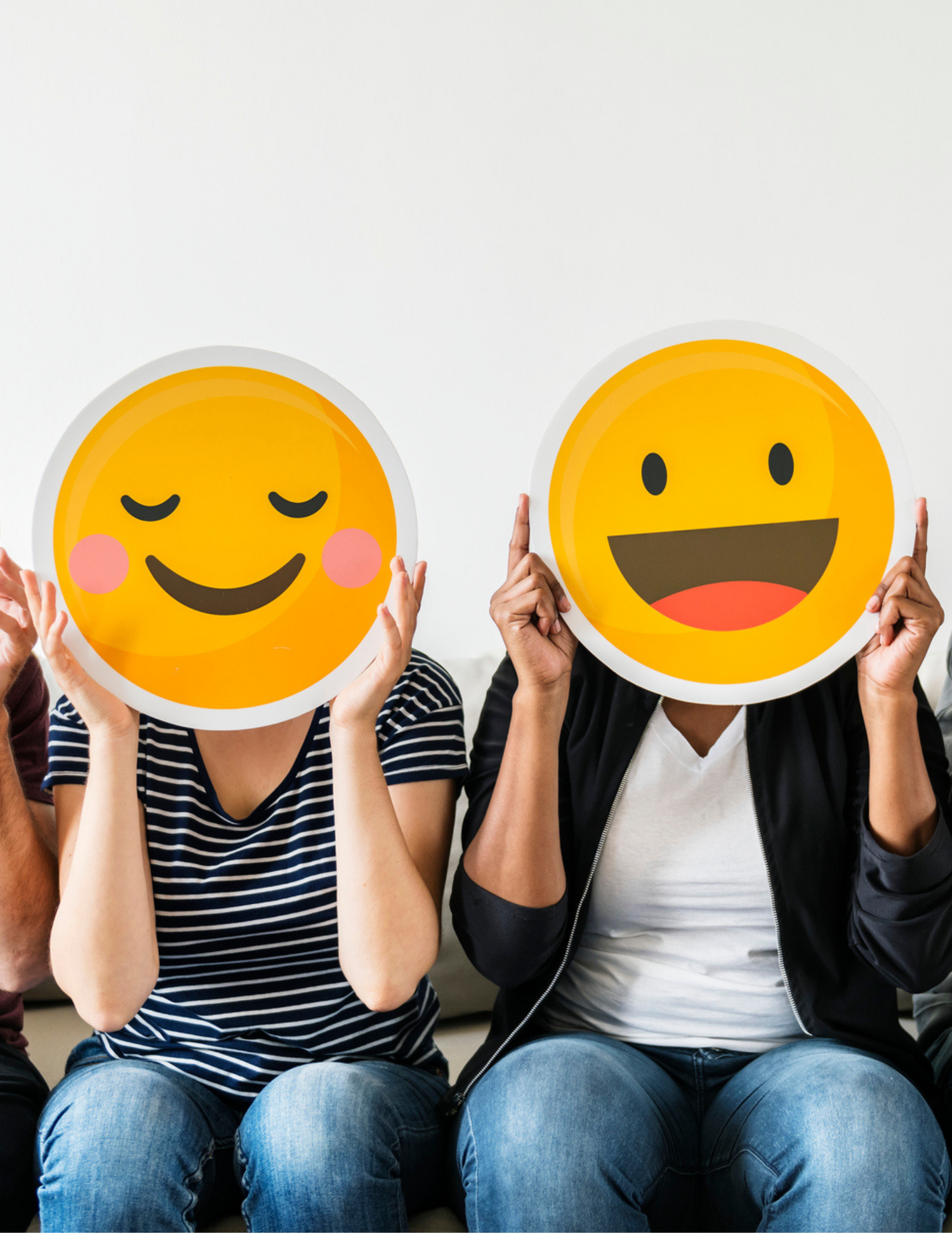 Two individuals sitting on a couch holding different smiling face emojis over their faces, having found their happy place.